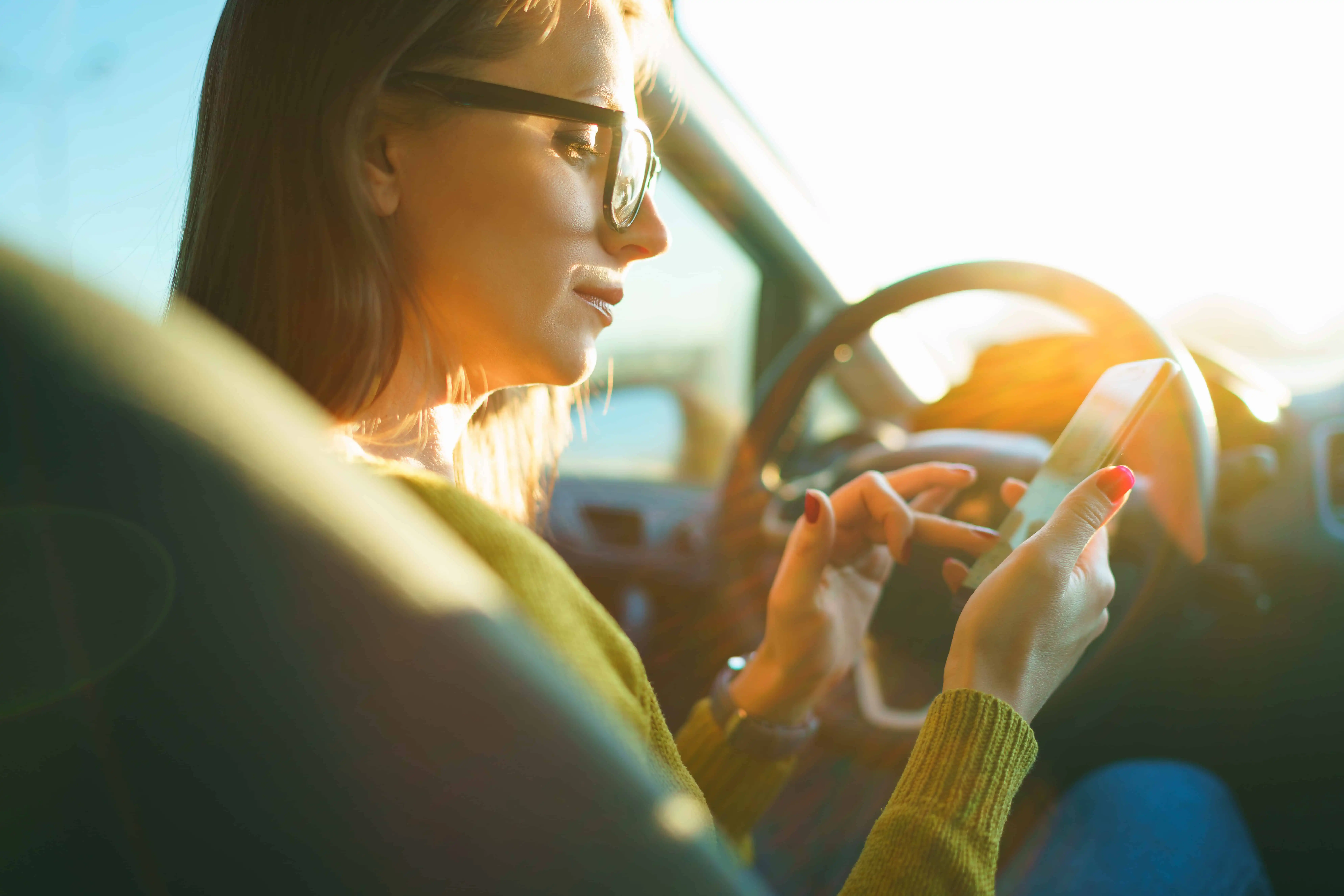 Happy woman in glasses uses a smartphone while driving a car at sunset